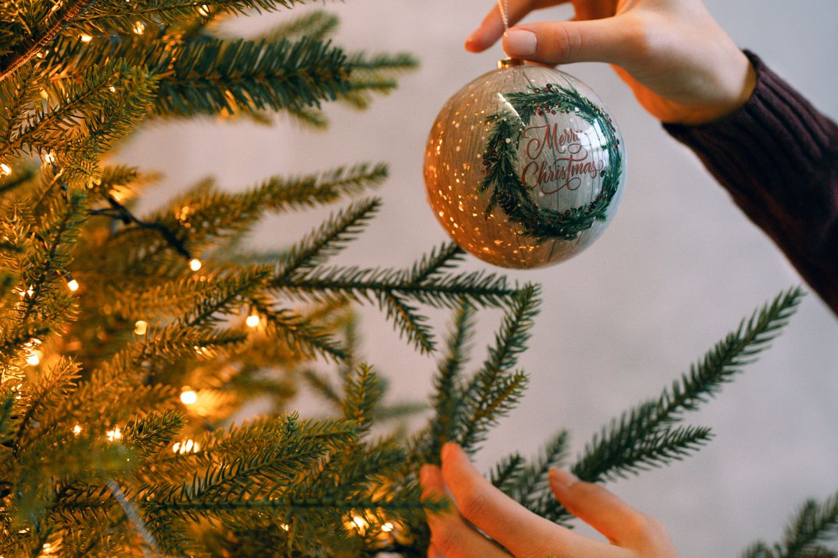 Gros plan sur une main qui accroche une boule de Noël dorée sur une branche de sapin décorée de guirlandes lumineuses
