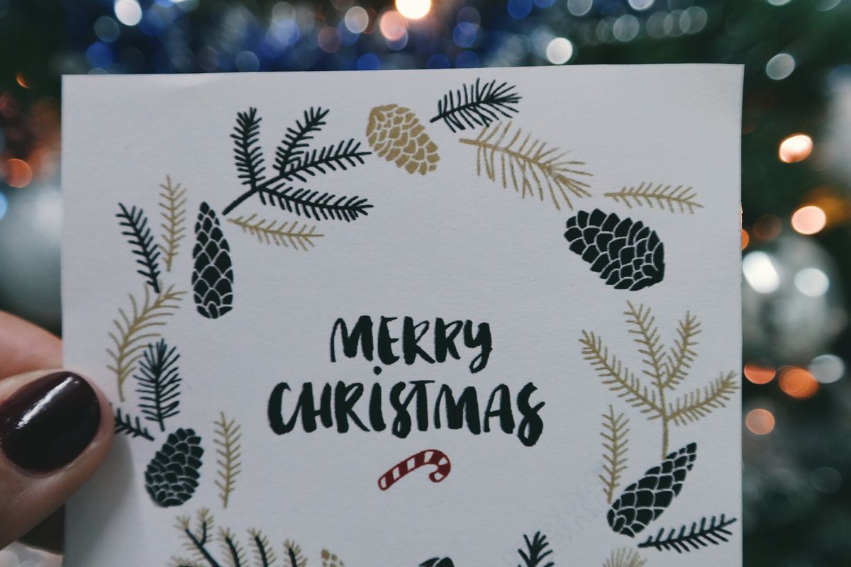 Une main tient une carte « Merry Christmas » ornée de branches de sapin, devant des lumières de Noël floues