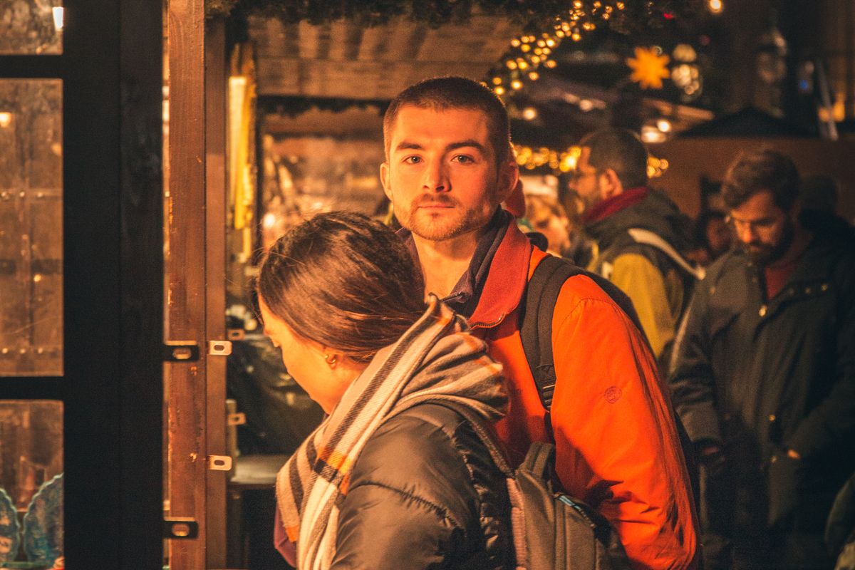 Une femme en écharpe et un homme en veste orange parmi les visiteurs d'un marché de Noël illuminé, de nuit, devant des étals décorés